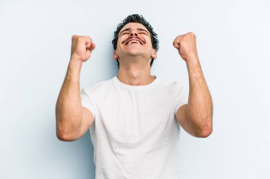 Mavi arka planda izole edilmiş genç beyaz bir adam zaferi, tutkuyu ve coşkuyu kutluyor, mutlu bir ifade..