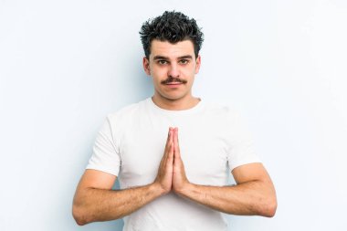 Kafkasyalı genç adam mavi arka planda izole edilmiş dua ediyor, bağlılık gösteriyor, dindar biri ilahi ilham arıyor..