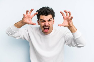 Mavi arka planda izole edilmiş genç beyaz adam pençelerini kedi taklidi yaparken gösteriyor, agresif bir hareket..