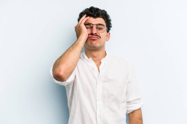 Kafkasyalı genç adam mavi arka planda izole edilmiş. Yorgun ve uykulu. Elini başının üstünde tutuyor..