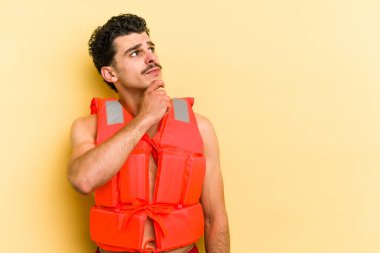 Young caucasian man wearing life jacket isolated on yellow background looking sideways with doubtful and skeptical expression.