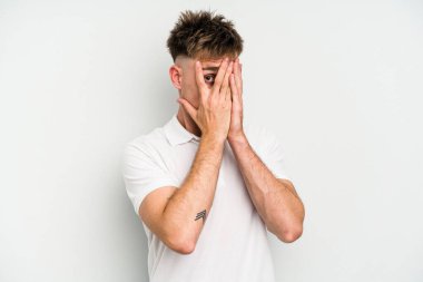 Beyaz arka planda izole edilmiş genç beyaz bir adam parmaklarının arasından korkmuş ve gergin bir şekilde göz kırpıyor..