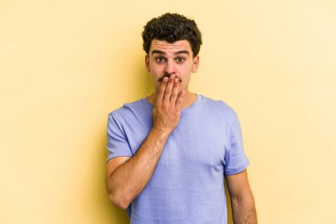 Sarı arka planda izole edilmiş genç beyaz adam şok olmuş, ağzı ellerle kaplanmış, yeni bir şey keşfetmeye hevesli..