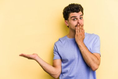 Sarı arka planda izole edilmiş genç beyaz adam, avuç içi fotokopi alanını elinde tutmaktan etkilendi..