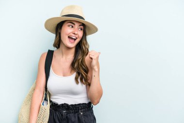 Young caucasian woman holding beach bag isolated on blue background points with thumb finger away, laughing and carefree.