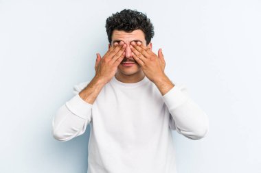 Mavi arka planda izole edilmiş genç beyaz adam elleriyle gözlerini kapatmaktan korkuyor..