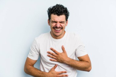 Mavi arka planda izole edilmiş genç beyaz adam mutlu bir şekilde gülüyor ve ellerini karnının üstünde tutarak eğleniyor..