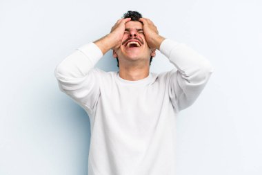 Mavi arka planda izole edilmiş genç beyaz adam ellerini başının üstünde tutarak neşe içinde gülüyor. Mutluluk kavramı.