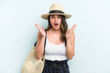 Young caucasian woman holding beach bag isolated on blue background surprised and shocked.