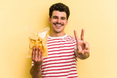 Young caucasian man holding a bag of chips isolated on yellow background joyful and carefree showing a peace symbol with fingers.