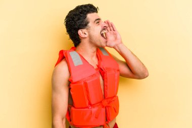 Young caucasian man wearing life jacket isolated on yellow background shouting and holding palm near opened mouth.