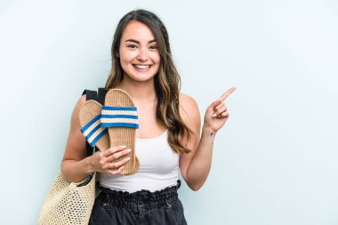 Young caucasian woman holding sandals isolated on blue background smiling and pointing aside, showing something at blank space.