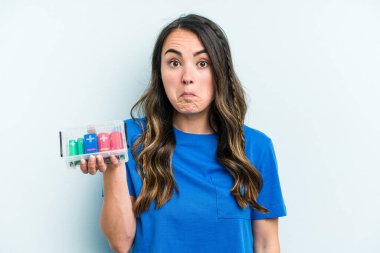 Young caucasian woman holding batteries isolated on blue background shrugs shoulders and open eyes confused.