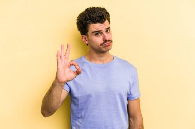 Sarı arka planda izole edilmiş genç beyaz bir adam bir göz kırptı ve elleriyle iyi bir jest yaptı..