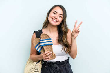 Young caucasian woman holding sandals isolated on blue background joyful and carefree showing a peace symbol with fingers.