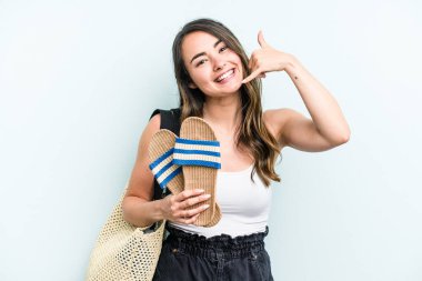 Young caucasian woman holding sandals isolated on blue background showing a mobile phone call gesture with fingers.