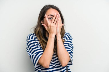 Beyaz arka planda izole edilmiş beyaz tenli genç bir kadın parmaklarının arasından korkmuş ve gergin bir şekilde göz kırpıyor..