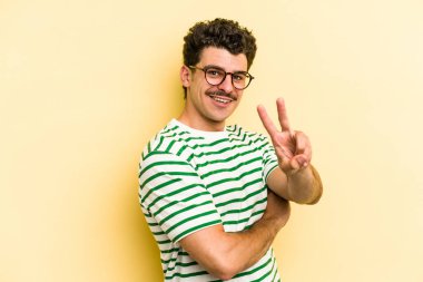 Sarı arka planda izole edilmiş genç beyaz adam zafer işareti gösteriyor ve gülümsüyor..