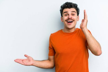 Mavi arka planda izole edilmiş genç beyaz bir adam avucunun içinde fotokopi alanı tutuyor, el üstünde tutuyor. Şaşırmış ve mutlu..