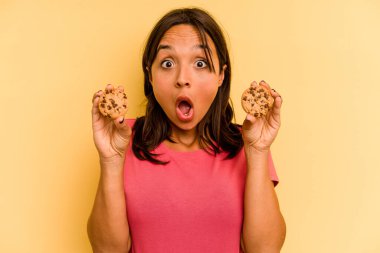 Young hispanic woman holding cookies isolated on yellow background