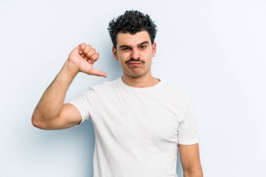 Mavi arka planda izole genç beyaz adam baş parmağını aşağı gösteriyor, hayal kırıklığı konsepti.