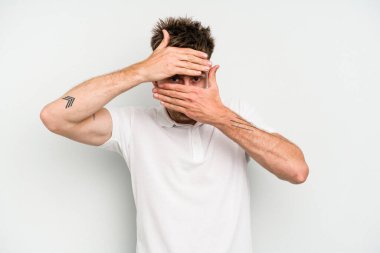 Beyaz arka planda izole edilmiş genç beyaz bir adam kameraya parmaklarıyla göz kırpıyor, yüzünü kapatmaktan utanıyor..