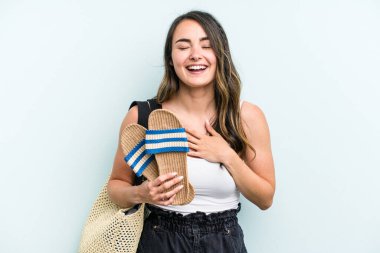 Young caucasian woman holding sandals isolated on blue background laughs out loudly keeping hand on chest.