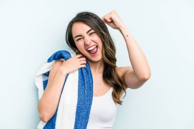 Young caucasian woman holding beach towel isolated on blue background raising fist after a victory, winner concept.