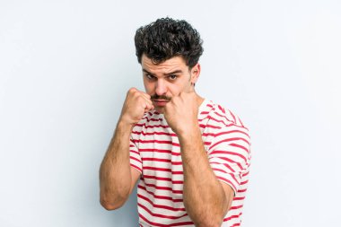 Kafkasyalı genç bir adam mavi arka planda izole edilmiş. Yumruk atıyor, sinirleniyor, kavga ediyor, kavga ediyor..