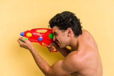Young caucasian man playing with a water gun isolated on yellow background