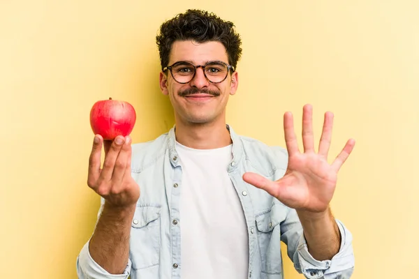 Young caucasian man holding an apple isolated on yellow background smiling cheerful showing number five with fingers.