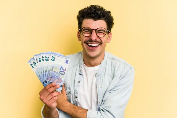 Young caucasian man holding banknotes isolated on yellow background laughing and having fun.