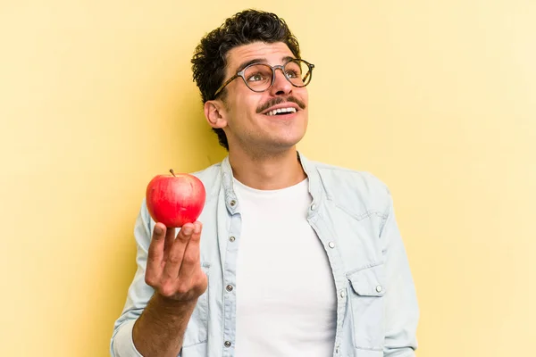 Young caucasian man holding an apple isolated on yellow background dreaming of achieving goals and purposes