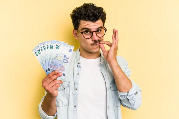 Young caucasian man holding banknotes isolated on yellow background with fingers on lips keeping a secret.