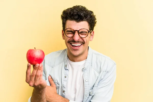 Young caucasian man holding an apple isolated on yellow background laughing and having fun.