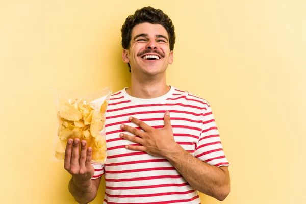 Young caucasian man holding a bag of chips isolated on yellow background laughs out loudly keeping hand on chest.