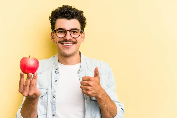 Young caucasian man holding an apple isolated on yellow background smiling and raising thumb up