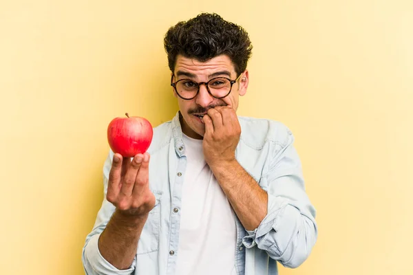 Young caucasian man holding an apple isolated on yellow background biting fingernails, nervous and very anxious.