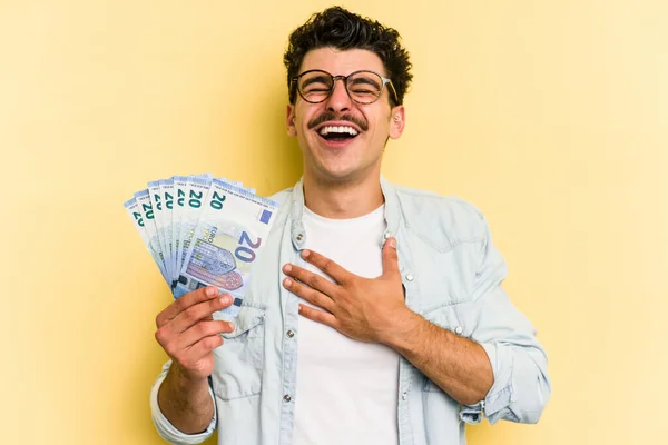 Young caucasian man holding banknotes isolated on yellow background laughs out loudly keeping hand on chest.