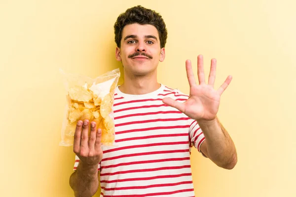 Young caucasian man holding a bag of chips isolated on yellow background smiling cheerful showing number five with fingers.