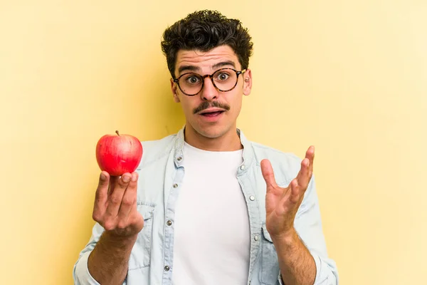 Young caucasian man holding an apple isolated on yellow background surprised and shocked.