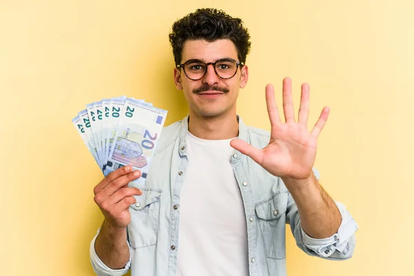 Young caucasian man holding banknotes isolated on yellow background smiling cheerful showing number five with fingers.