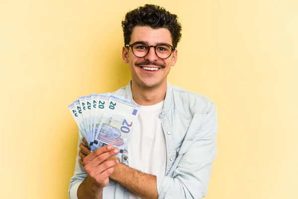 Young caucasian man holding banknotes isolated on yellow background happy, smiling and cheerful.