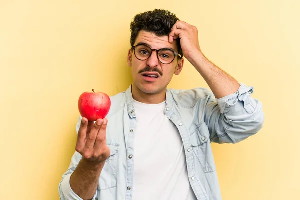 Young caucasian man holding an apple isolated on yellow background being shocked, she has remembered important meeting.