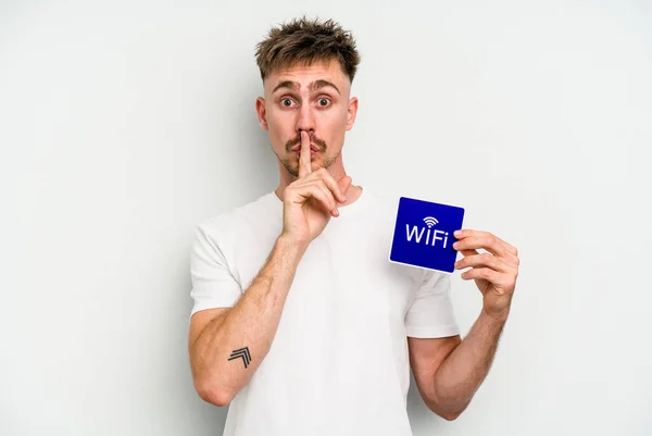 Young caucasian man holding wifi placard isolated on white background keeping a secret or asking for silence.