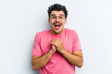 Kafkasyalı genç adam mavi arka planda izole edilmiş şans için dua ediyor, şaşırmış ve ağzını açıp önüne bakıyor..