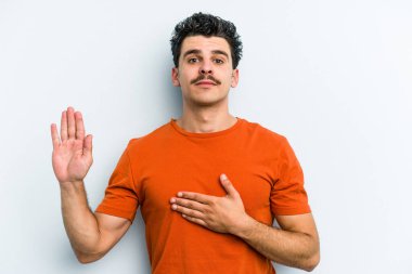 Kafkasyalı genç adam mavi arka planda izole edilmiş yemin ediyor, elini göğsüne koyuyor..