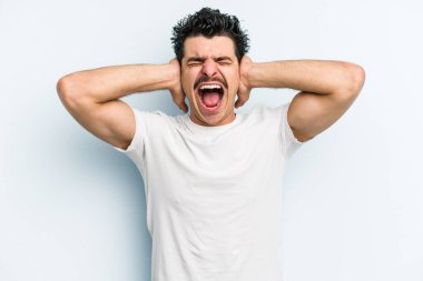 Kafkasyalı genç adam mavi arka planda izole edilmiş kulakları kulaklarıyla kapatıyor çok fazla ses duymamaya çalışıyor..