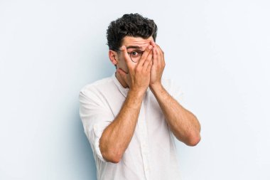 Kafkasyalı genç adam mavi arka planda izole edilmiş. Korkmuş ve gergin parmaklarıyla göz kırp..