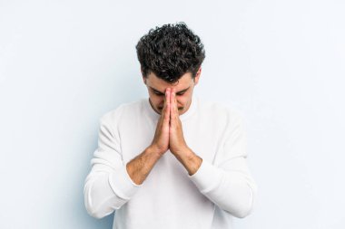 Kafkasyalı genç adam mavi arka planda izole edilmiş dua ediyor, bağlılık gösteriyor, dindar biri ilahi ilham arıyor..